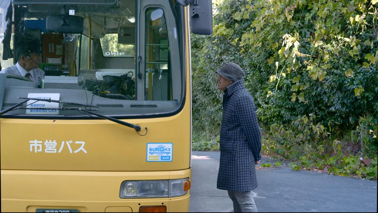 Ein gelber Bus hält auf der Straße und hat die Fahrertür geöffnet. Ein Mann steht vor dem Bus und spricht mit dem Fahrer.
