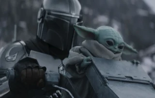 Szenenbild aus The Mandalorian and Grogu: Eine gepanzerte Figur mit glänzendem Helm hält ein metallisches Objekt, während eine kleine, grünhäutige Kreatur mit großen Ohren und einem beigefarbenen Umhang daneben sitzt. [erstellt mit KI]