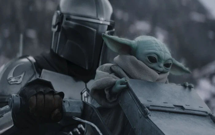 Szenenbild aus The Mandalorian and Grogu: Eine gepanzerte Figur mit glänzendem Helm hält ein metallisches Objekt, während eine kleine, grünhäutige Kreatur mit großen Ohren und einem beigefarbenen Umhang daneben sitzt. [erstellt mit KI]
