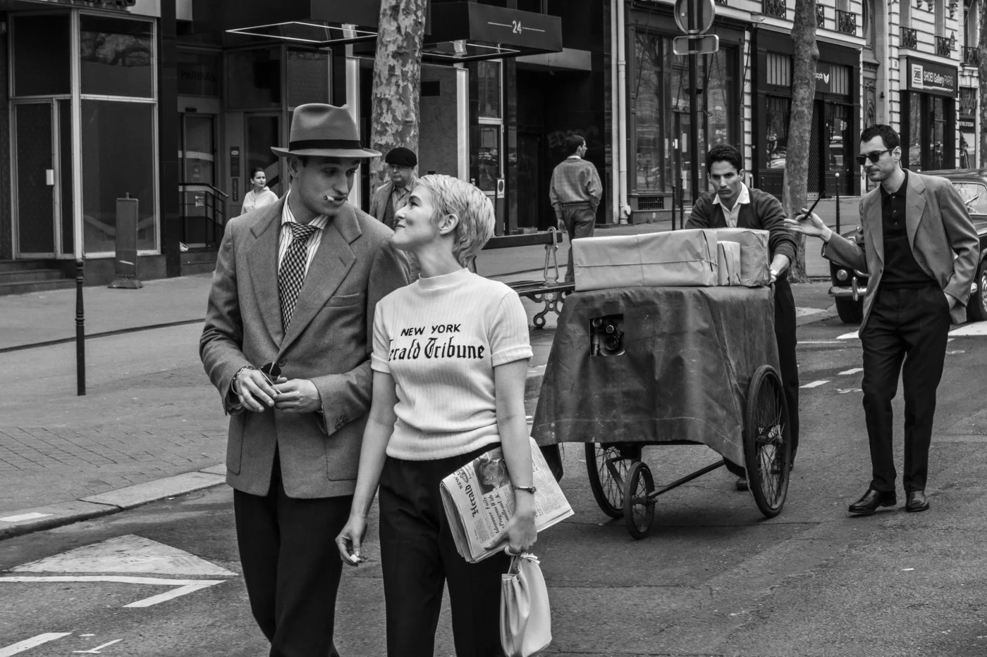 Szenenbild aus "Nouvelle VagueI": Im Vordergrund gehen zwei Personen nebeneinander auf einer Straße. Die Person rechts trägt ein helles Oberteil mit dem Schriftzug 'NEW YORK Herald Tribune' und hält eine Zeitung in der Hand. Die Person links trägt einen Anzug und einen Hut. Im Hintergrund schiebt eine weitere Person einen großen Wagen mit Paketen, während eine weitere Person in dunkler Kleidung und Sonnenbrille hinterherläuft. Die Szene spielt in einer städtischen Umgebung mit Gebäuden, Schaufenstern und geparkten Autos.