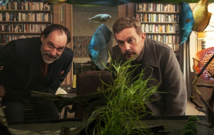 Szenenbild aus Die Ältern: Zwei Männer mit Bart und ernster Miene beugen sich nach vorne und blicken durch ein großes Aquarium. Mehrere bunte Fische schwimmen im Wasser, im Vordergrund sind grüne Wasserpflanzen und Dekorationen zu sehen. Im Hintergrund steht ein großes Bücherregal mit vielen Büchern. [erstellt mit KI]
