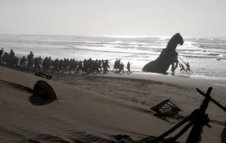 Am Strand ziehen zahlreiche Menschen mit Seilen eine große Pferdstatue aus dem Wasser. Im Vordergrund liegen verstreute Holzteile und Netze im Sand. Die Brandung und das Meer sind im Hintergrund sichtbar, während die Sonne tief steht und lange Schatten wirft. [erstellt mit KI]