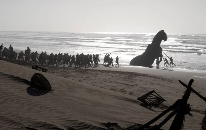 Am Strand ziehen zahlreiche Menschen mit Seilen eine große Pferdstatue aus dem Wasser. Im Vordergrund liegen verstreute Holzteile und Netze im Sand. Die Brandung und das Meer sind im Hintergrund sichtbar, während die Sonne tief steht und lange Schatten wirft. [erstellt mit KI]