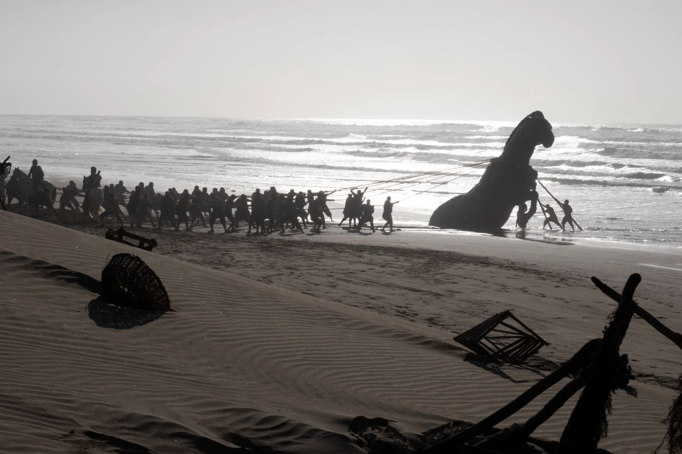 Am Strand ziehen zahlreiche Menschen mit Seilen eine große Pferdstatue aus dem Wasser. Im Vordergrund liegen verstreute Holzteile und Netze im Sand. Die Brandung und das Meer sind im Hintergrund sichtbar, während die Sonne tief steht und lange Schatten wirft. [erstellt mit KI]