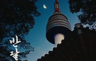 Szenenbild aus Ban Dal: Im Vordergrund ragt das Dach eines traditionellen koreanischen Gebäudes schräg ins Bild. Dahinter erhebt sich ein hoher Turm mit rundem Aussichtsbereich und Antenne, der von unten nach oben fotografiert wurde. Der Himmel ist dunkelblau, im oberen Bildbereich steht eine schmale Mondsichel. Um den Turm und das Dach sind die Silhouetten von Bäumen zu sehen. Links unten sind große, weiße Schriftzeichen in koreanischer Sprache zu erkennen. [erstellt mit KI]