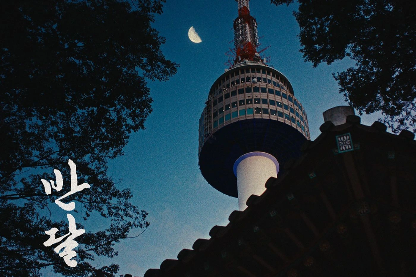 Szenenbild aus Ban Dal: Im Vordergrund ragt das Dach eines traditionellen koreanischen Gebäudes schräg ins Bild. Dahinter erhebt sich ein hoher Turm mit rundem Aussichtsbereich und Antenne, der von unten nach oben fotografiert wurde. Der Himmel ist dunkelblau, im oberen Bildbereich steht eine schmale Mondsichel. Um den Turm und das Dach sind die Silhouetten von Bäumen zu sehen. Links unten sind große, weiße Schriftzeichen in koreanischer Sprache zu erkennen. [erstellt mit KI]
