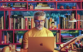 Szenenbild aus Bitteres Fest: Ein Mann mit grauem Haar und Bart sitzt an einem Schreibtisch vor einem aufgeklappten Laptop. Er trägt ein gelbes Oberteil und eine Brille mit dunklem Rahmen. Im Hintergrund steht ein Regal mit zahlreichen Büchern und einigen dekorativen Gegenständen. Die Bücher sind in mehreren Reihen angeordnet, einige Titel sind deutlich lesbar. Die Szene ist farbenfroh und wirkt durch die Beleuchtung lebendig. [erstellt mit KI]