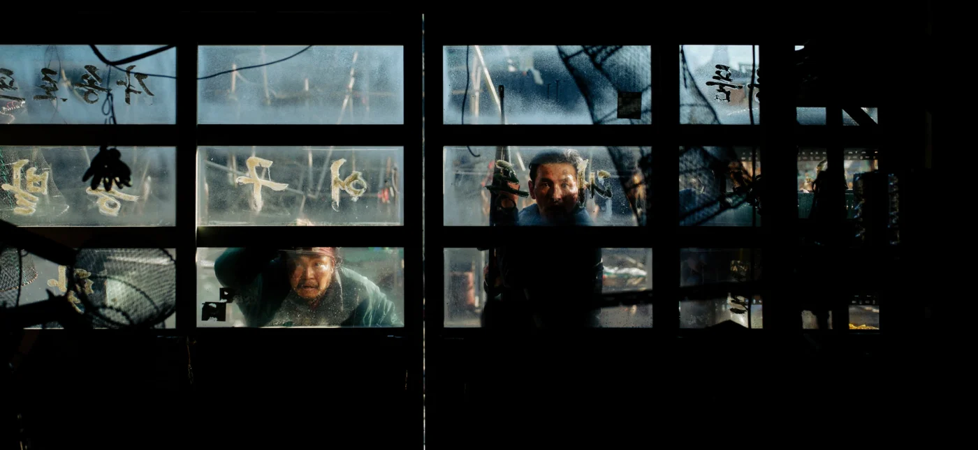 Szenenbild aus Hope: Zwei Personen sind durch die Scheiben eines großen, in mehrere Felder unterteilten Fensters zu sehen. Die Scheiben sind von außen beschlagen und teilweise mit Zeichnungen oder Symbolen versehen. [erstellt mit KI]