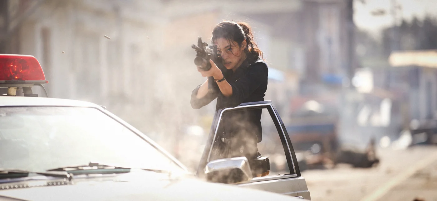 Szenenbild aus Hope: Eine Frau mit ernstem Gesichtsausdruck steht hinter einer geöffneten Autotür eines weißen Fahrzeugs mit eingeschaltetem Blaulicht. Sie hält ein Gewehr im Anschlag und zielt in Richtung des Bildvordergrunds. Staub und Rauchpartikel liegen in der Luft, im Hintergrund sind unscharfe Gebäude und weitere Fahrzeuge zu erkennen. Die Szene wirkt angespannt und dynamisch. [erstellt mit KI]