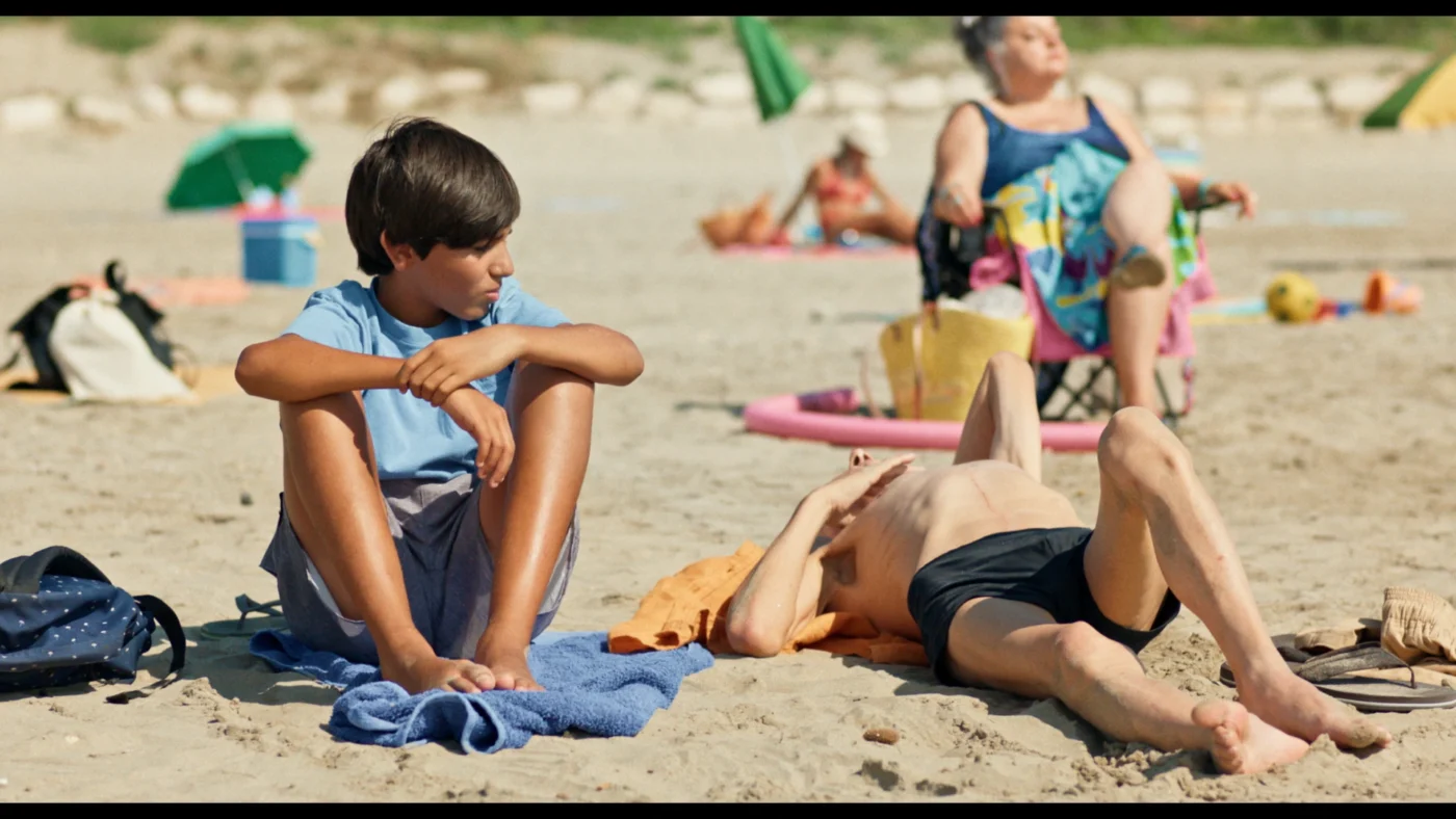 Szenenbild aus Pale Sun: Ein Junge mit braunen Haaren sitzt auf einem blauen Handtuch am Sandstrand. Er trägt ein hellblaues T-Shirt und kurze Hosen und blickt auf eine Person, die neben ihm auf dem Rücken liegt. Die liegende Person ist oberkörperfrei und hat einen Arm über die Stirn gelegt. Im Hintergrund sind weitere Menschen auf bunten Handtüchern und Liegen zu sehen, einige mit Sonnenschirmen. Die Szene spielt bei sonnigem Wetter.
