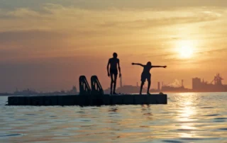 Zwei Personen stehen auf einem Steg, der von Wasser umgeben ist. Die Sonne steht tief am Himmel und taucht die Szene in warmes Licht. Im Hintergrund sind Industrieanlagen und Schornsteine zu erkennen. Die Wasseroberfläche reflektiert die Farben des Sonnenuntergangs. Eine Person steht aufrecht, die andere breitet die Arme aus. [erstellt mit KI]