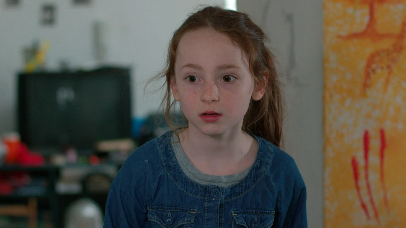 Szenenbild aus dem Film "Zoey & Ben" Ein Mädchen mit rotbraunen Haaren und dunkelblauem Oberteil steht in einem Wohnzimmer und schaut erschrocken an der Kamera vorbei.