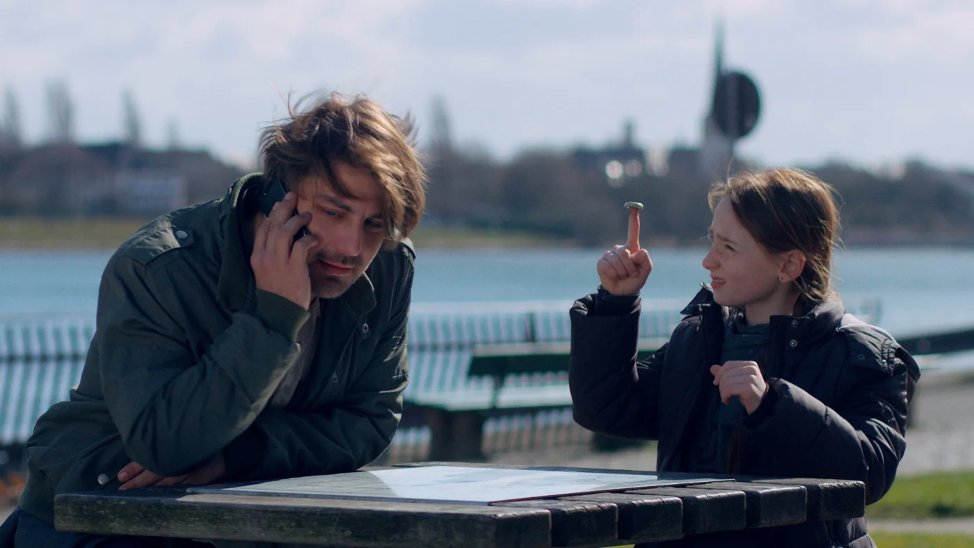 Szenenbild aus Zoey & Ben: Ein Mann und ein Mädchen sitzen an einem Holztisch im Freien. Im Hintergrund ist ein Fluss zu sehen. Der Mann telefoniert an seinem Handy, während das Mädchen neben ihm einen Kronkorken auf ihrem rechten Zeigefinger balanciert.